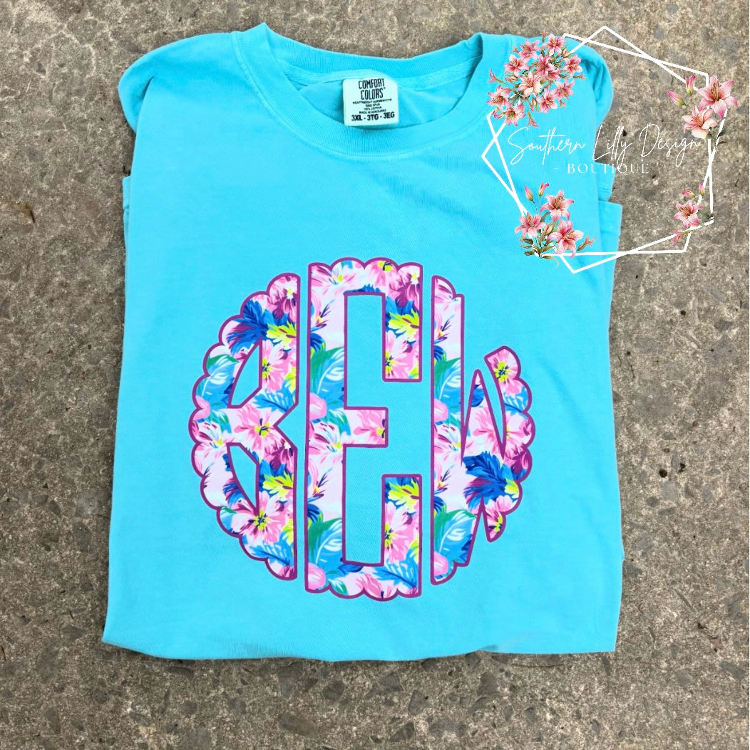 Light blue t-shirt with floral monogram design on a concrete surface