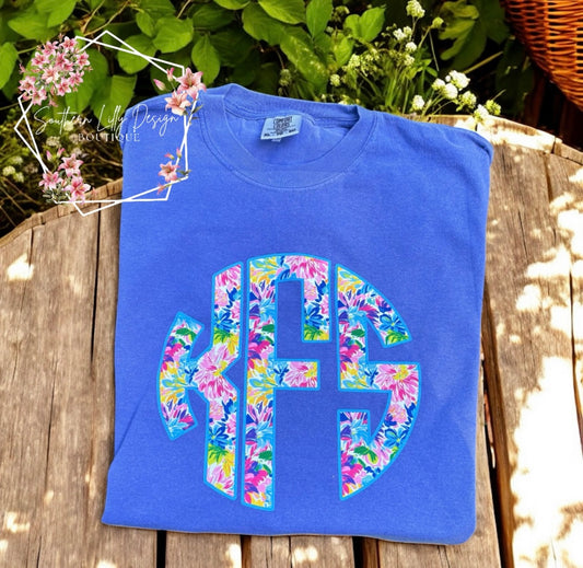 Blue t-shirt with floral monogram on a wooden surface with flowers in the background